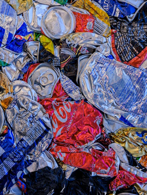 Three large black plastic rubbish bags and a cardboard box, all filled with waste material, are placed on the curbside pavement in front of a tall, dark wooden fence and green foliage. The rubbish bag
