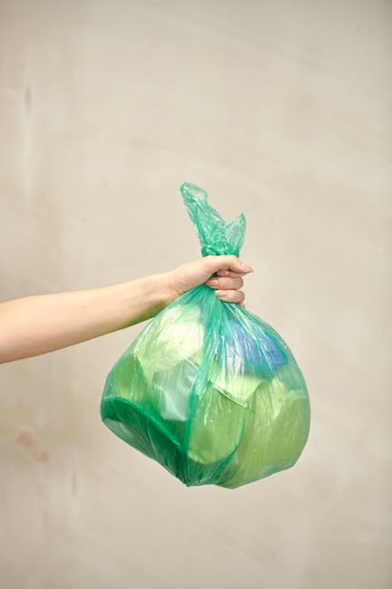A person's arm extends from the left side of the image, holding a transparent green plastic bag filled with various types of household rubbish. The bag appears to be loosely gathered at the top and ti