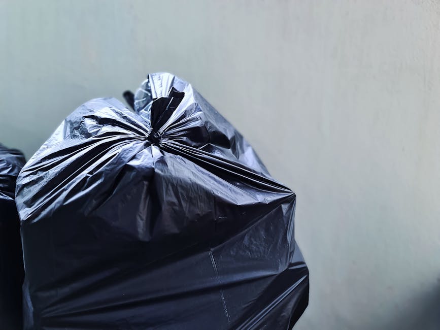 A person's arm extends from the left side of the image, holding a transparent green plastic bag filled with various types of household rubbish. The bag appears to be loosely gathered at the top and ti