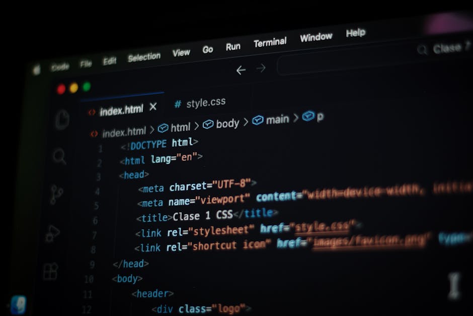 A person seated at a desk working on a silver laptop, which displays a screen filled with colorful lines of code indicating programming or software development. The individual is wearing a light blue 